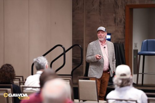 Presenter in a gray blazer and pink shirt speaks to an audience in a conference room, wearing a cap and wireless mic.