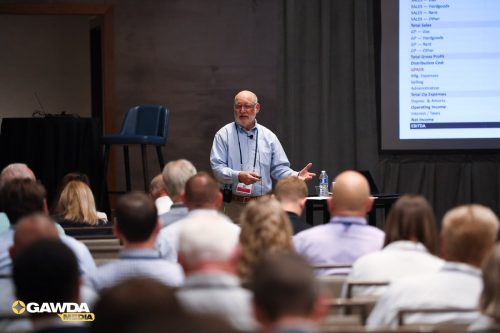 Older man in a light blue shirt explains a slide to a seated audience; projector screen with data on the right, water bottle on the table, GAWDA Media logo bottom left