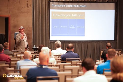 Presenter in a blazer and cap speaks to an audience in a conference room with a slide titled 'How do you bill rent' on the screen behind him, showing rent categories like Daily, Monthly, peak monthly, and demurrage.
