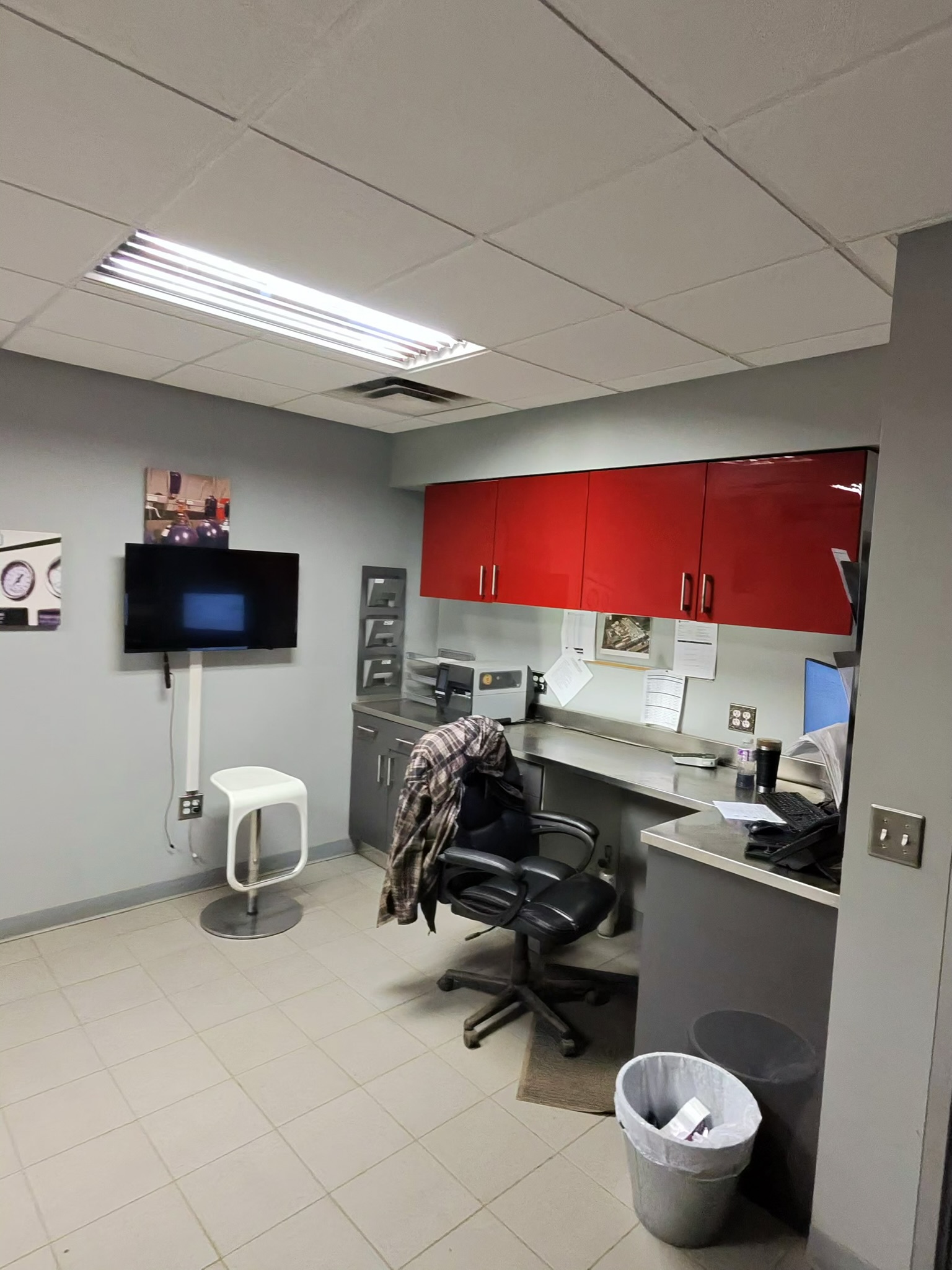 Office break room with red wall cabinets above a stainless counter, desk chair, computer, and trash can nearby.