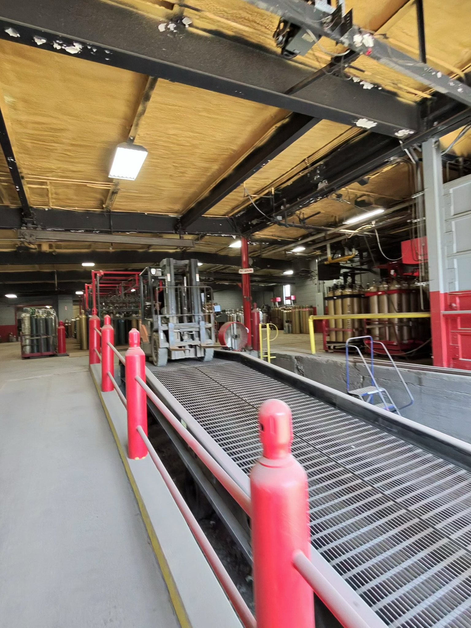 Industrial interior with grated floor trench and red safety posts along a walkway, heavy machinery in the background.