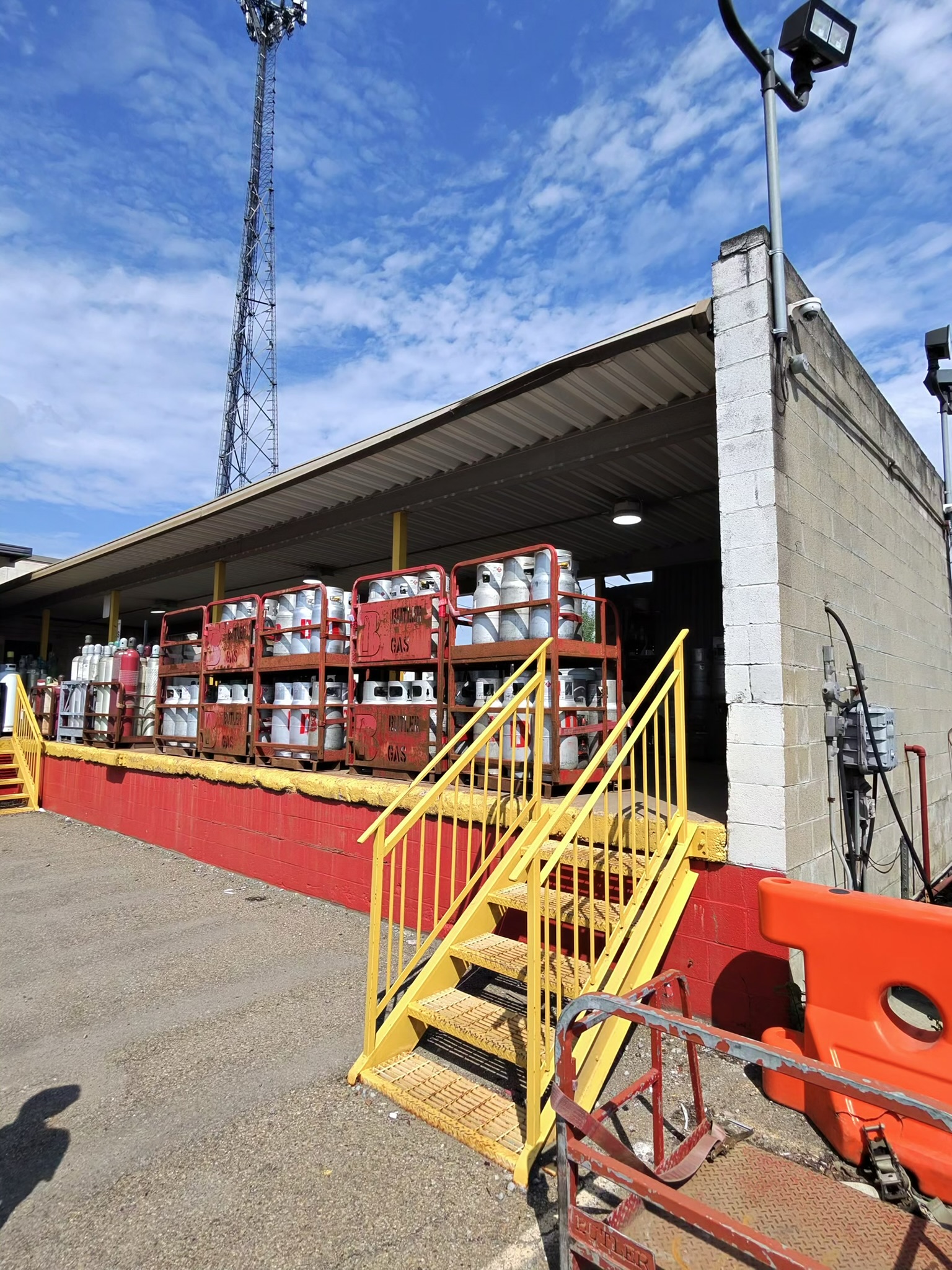 Outdoor propane cylinder storage on red pallets under a metal roof, with yellow stairs and a concrete block building nearby.