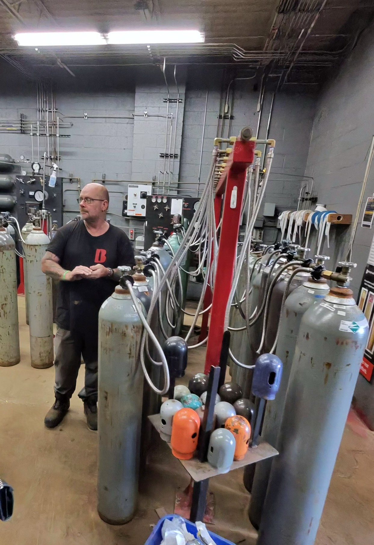 Man in a black t‑shirt standing among tall gas cylinders and tangled hoses in an industrial lab setting.