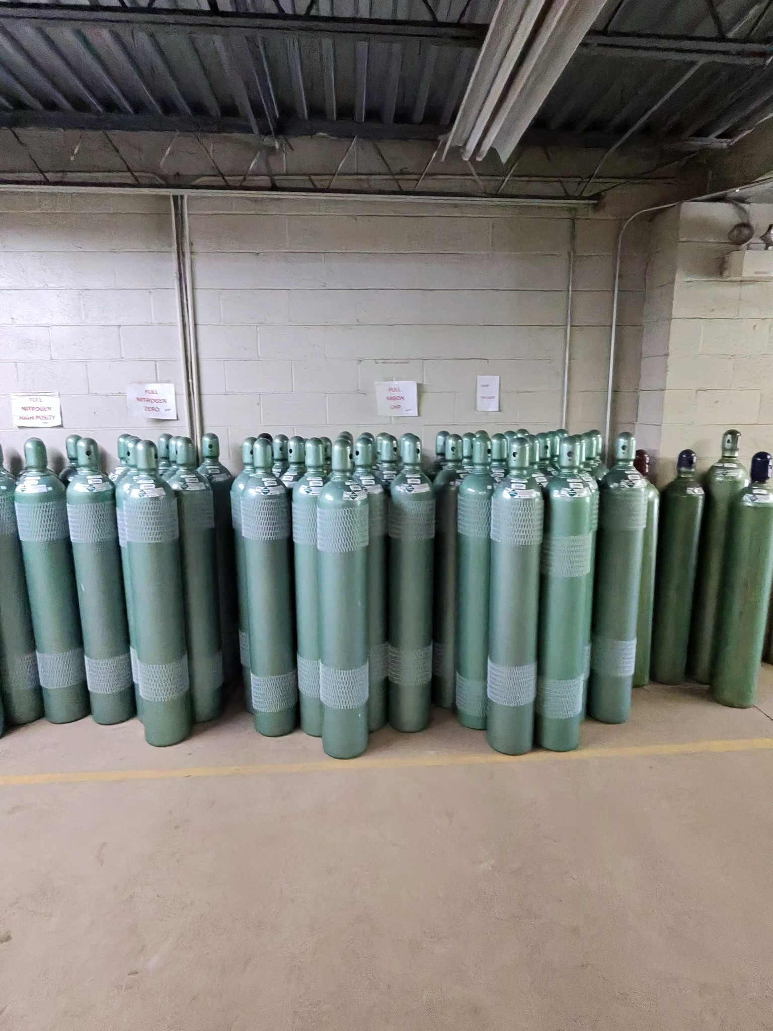 Green compressed gas cylinders lined up against a wall in a warehouse, with signs posted above them and exposed ceiling beams behind.