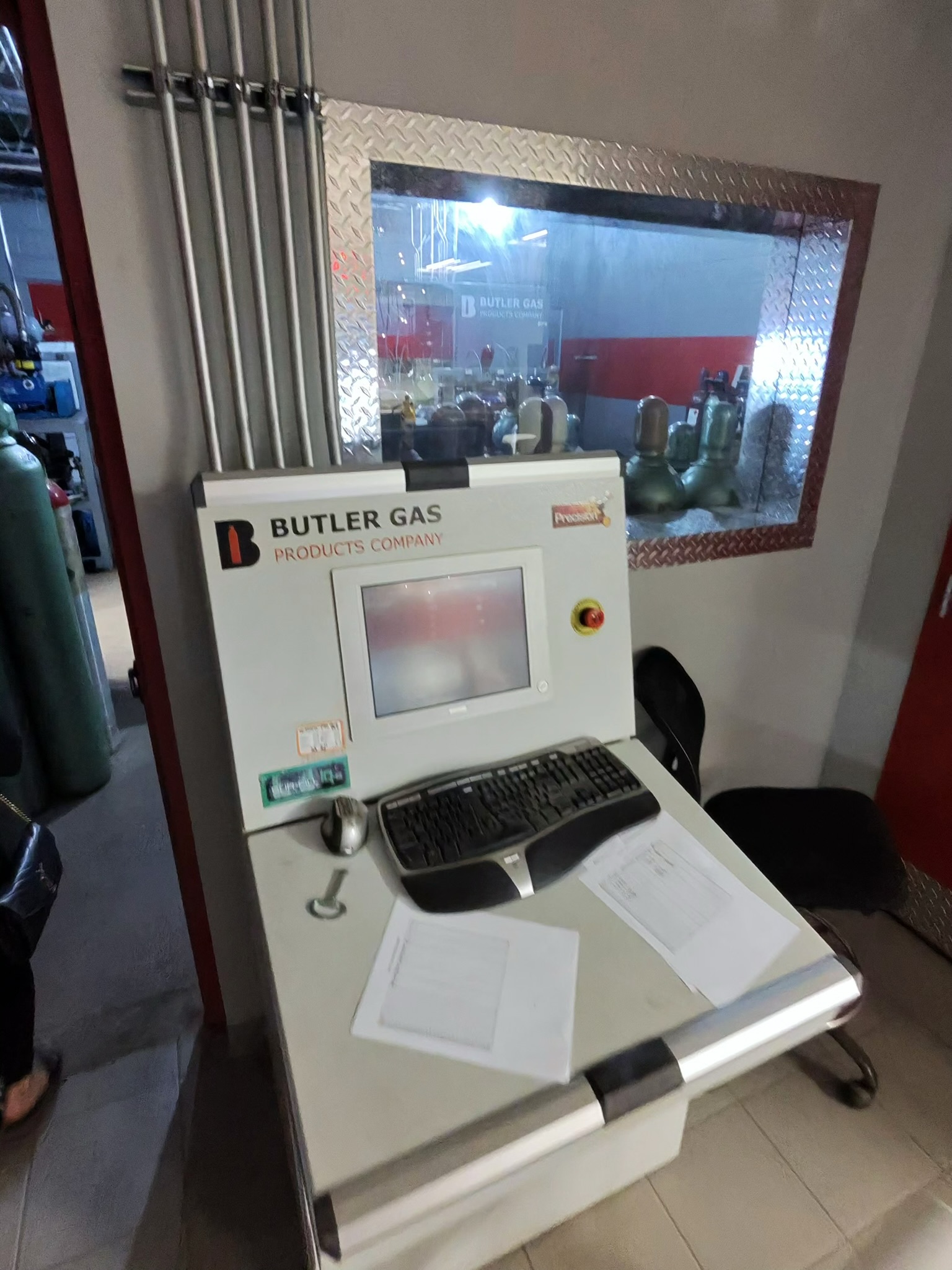 Industrial control terminal labeled 'BUTLER GAS PRODUCTS COMPANY' with keyboard and mouse on a counter, overlooking a room of gas bottles through a diamond-plate window
