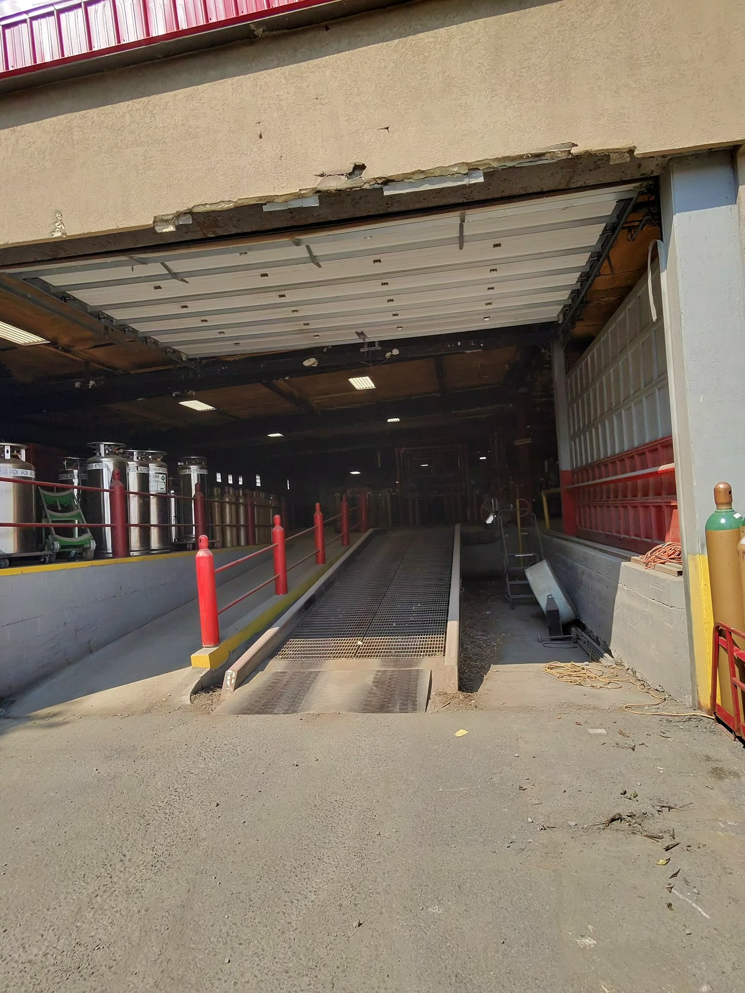 Industrial loading dock with an open roll-up door, metal ramp, and red safety rails along a concrete entryway. Left side shows gas cylinders nearby.