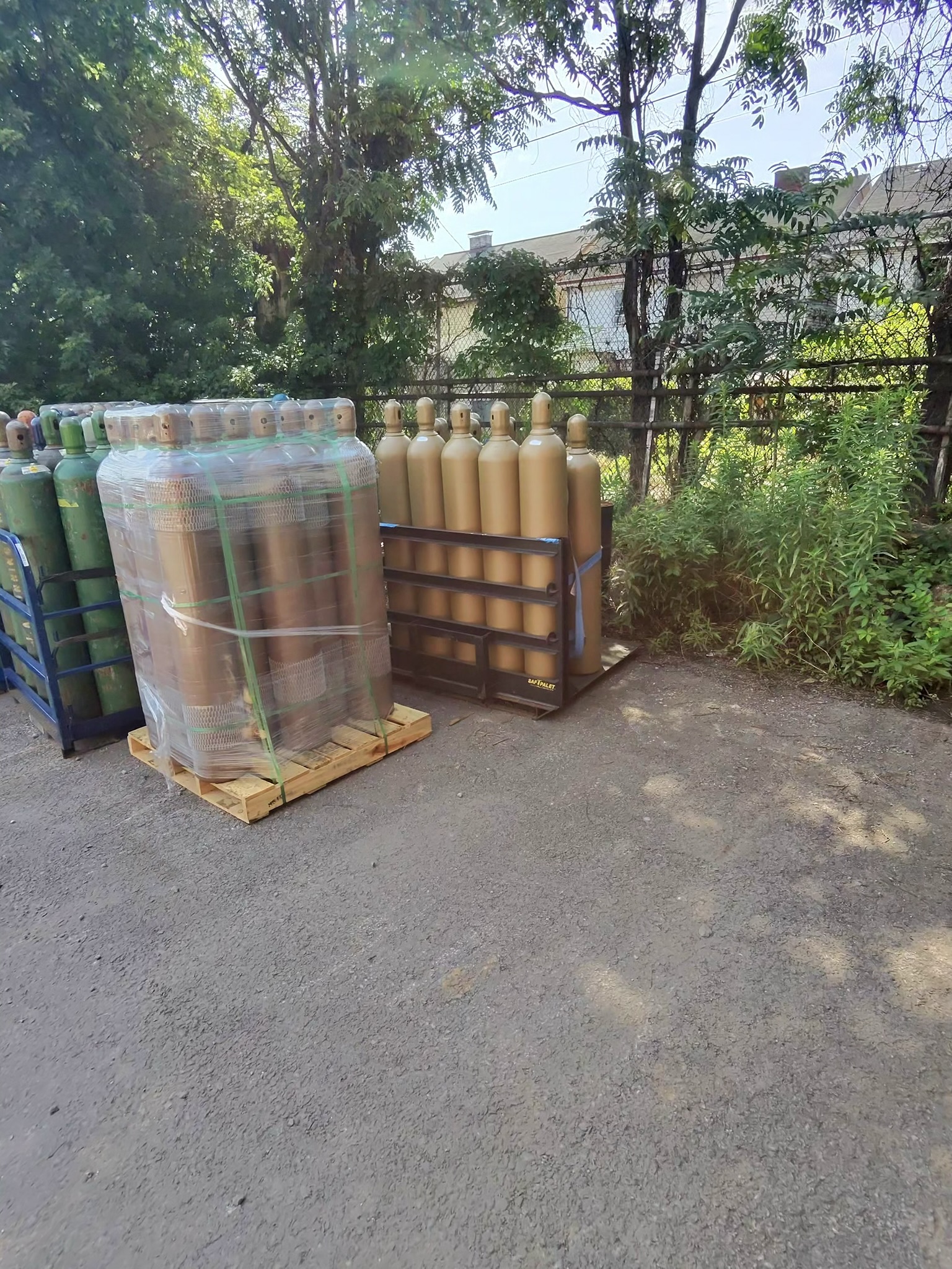 Row of beige gas cylinders on a metal rack outside, with a chain-link fence and trees in the background.