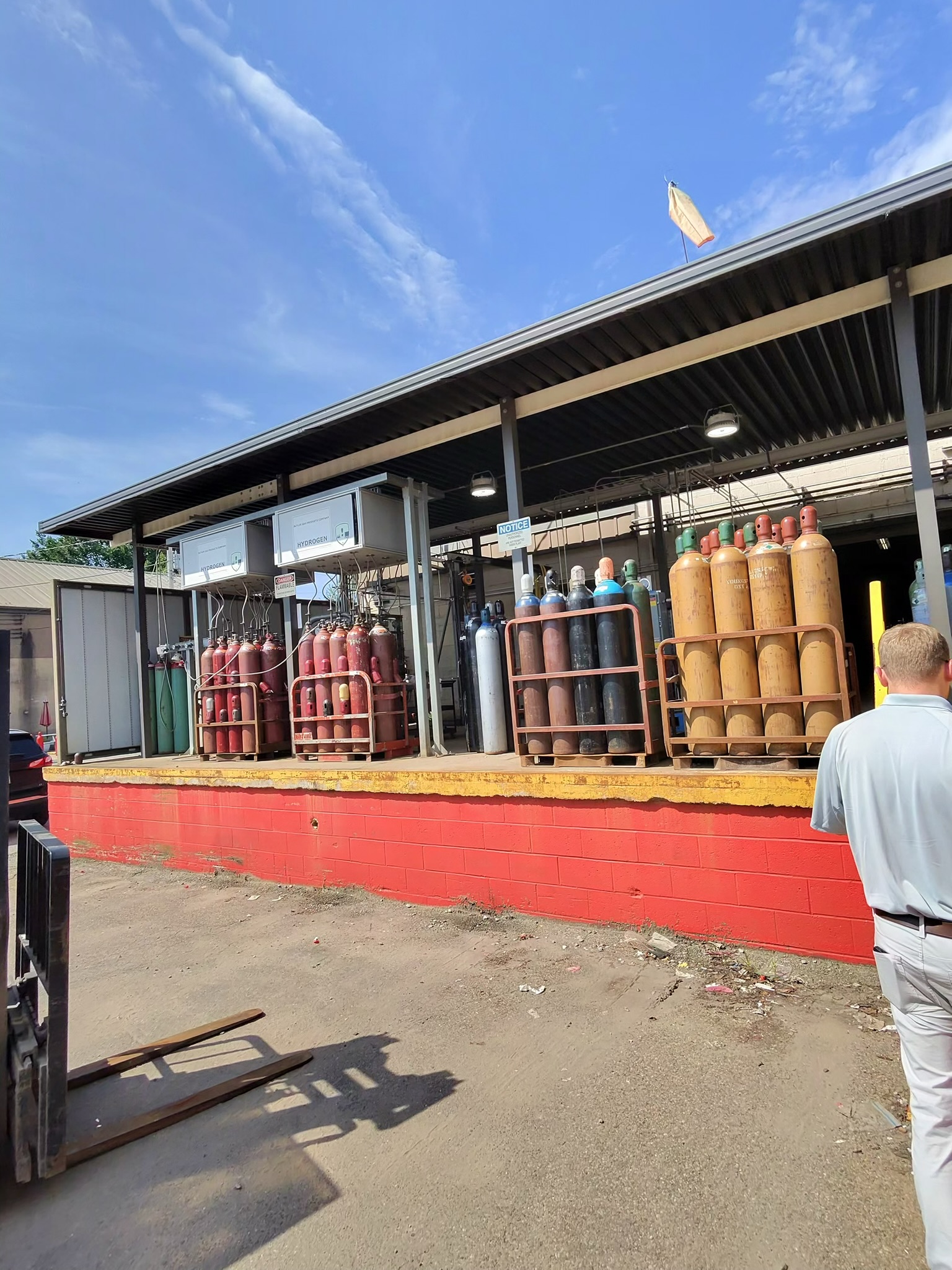 Outdoor industrial gas storage area with tall colored cylinders on racks under a metal roof; a red barrier wall in the foreground and a person partly visible on the right.