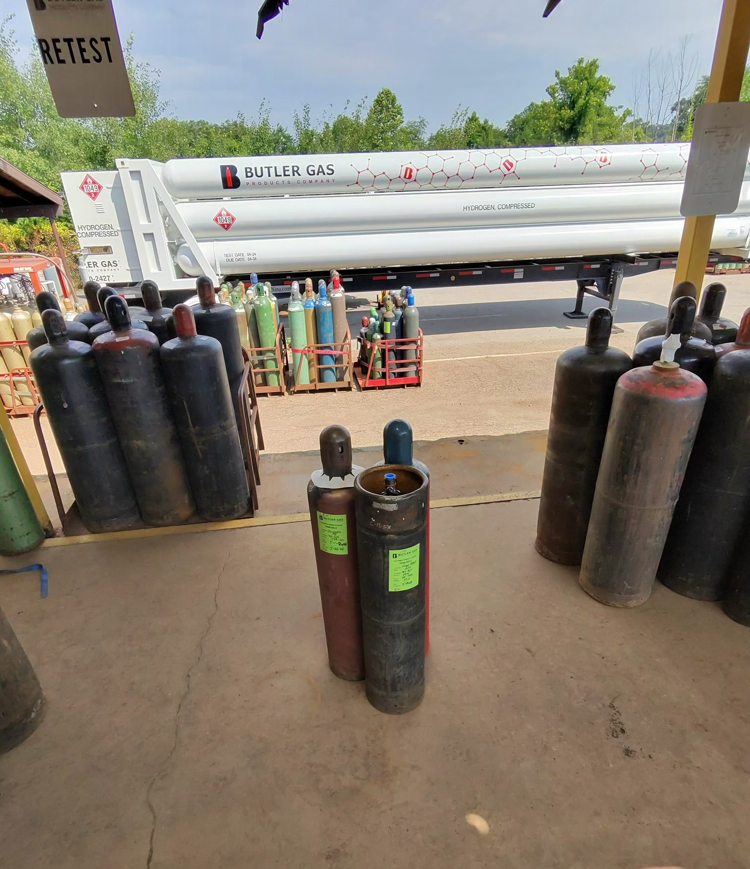 Row of tall black gas cylinders in the foreground with a large white hydrogen tanker trailer labeled Butler Gas in the background.