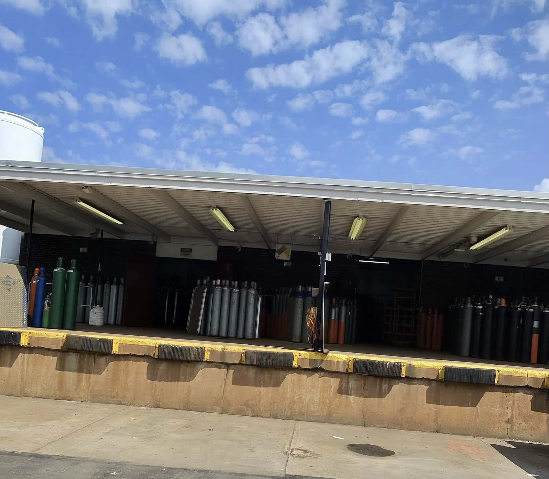 Open-front industrial warehouse with rows of gas cylinders lined up under a corrugated metal roof; a blue sky with scattered clouds overhead and a yellow loading dock curb in the foreground.