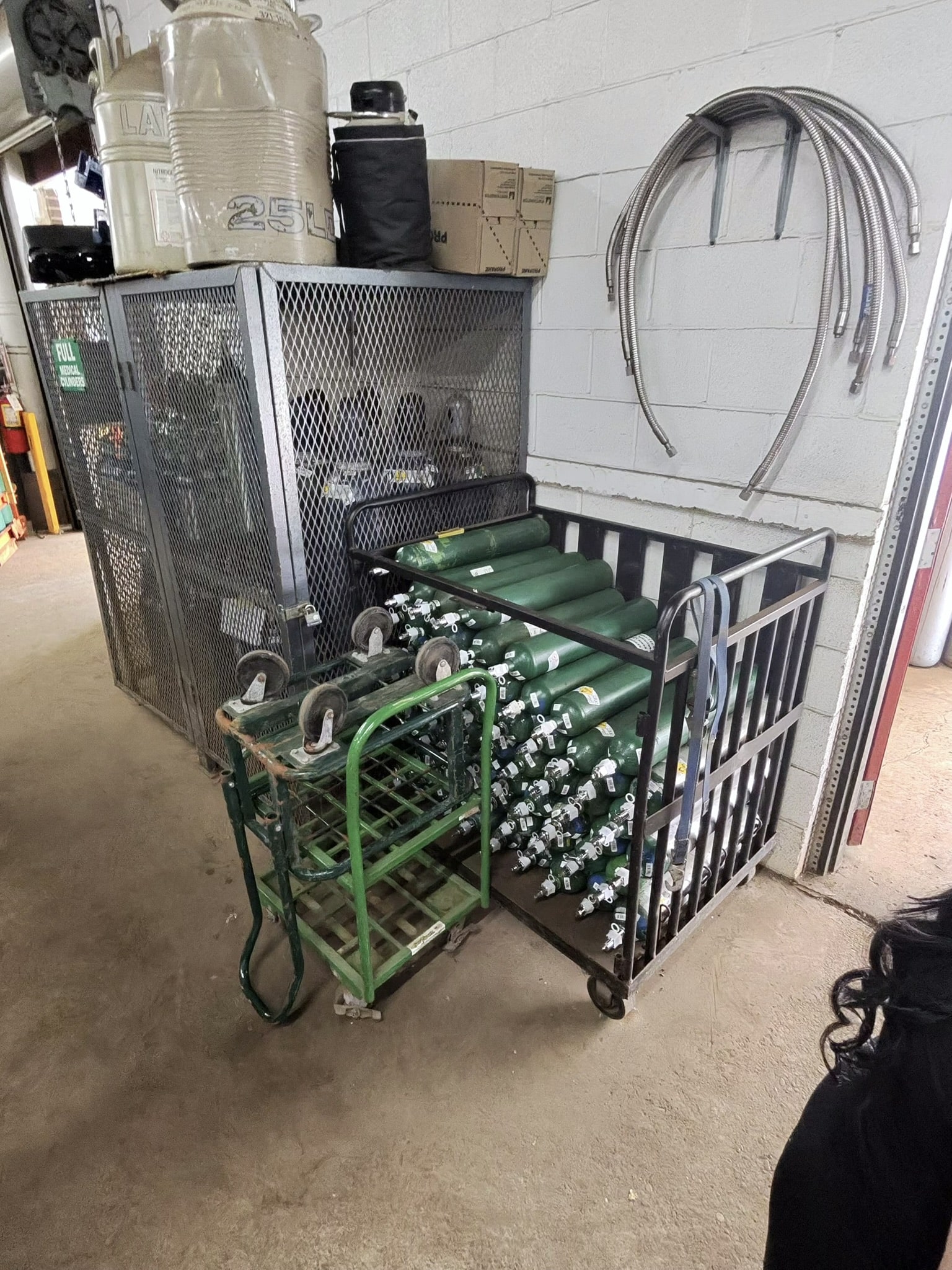 Industrial utility area with gas cylinders on a green cart next to a metal cage and coiled hoses on the wall