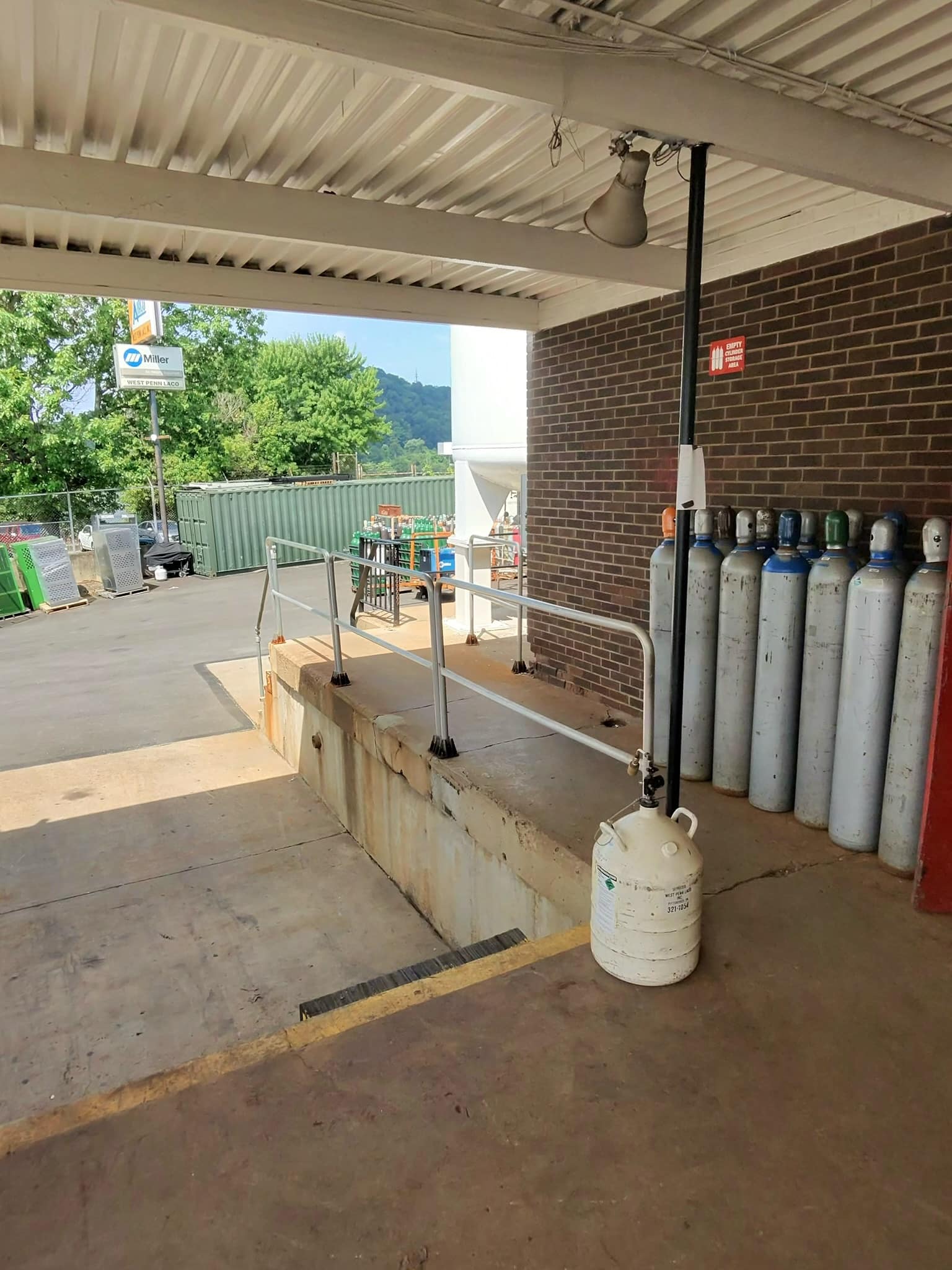 Loading dock area with a row of tall propane cylinders lined up along a brick wall and a portable gas cylinder in the foreground, railing and outdoor equipment visible in the background.