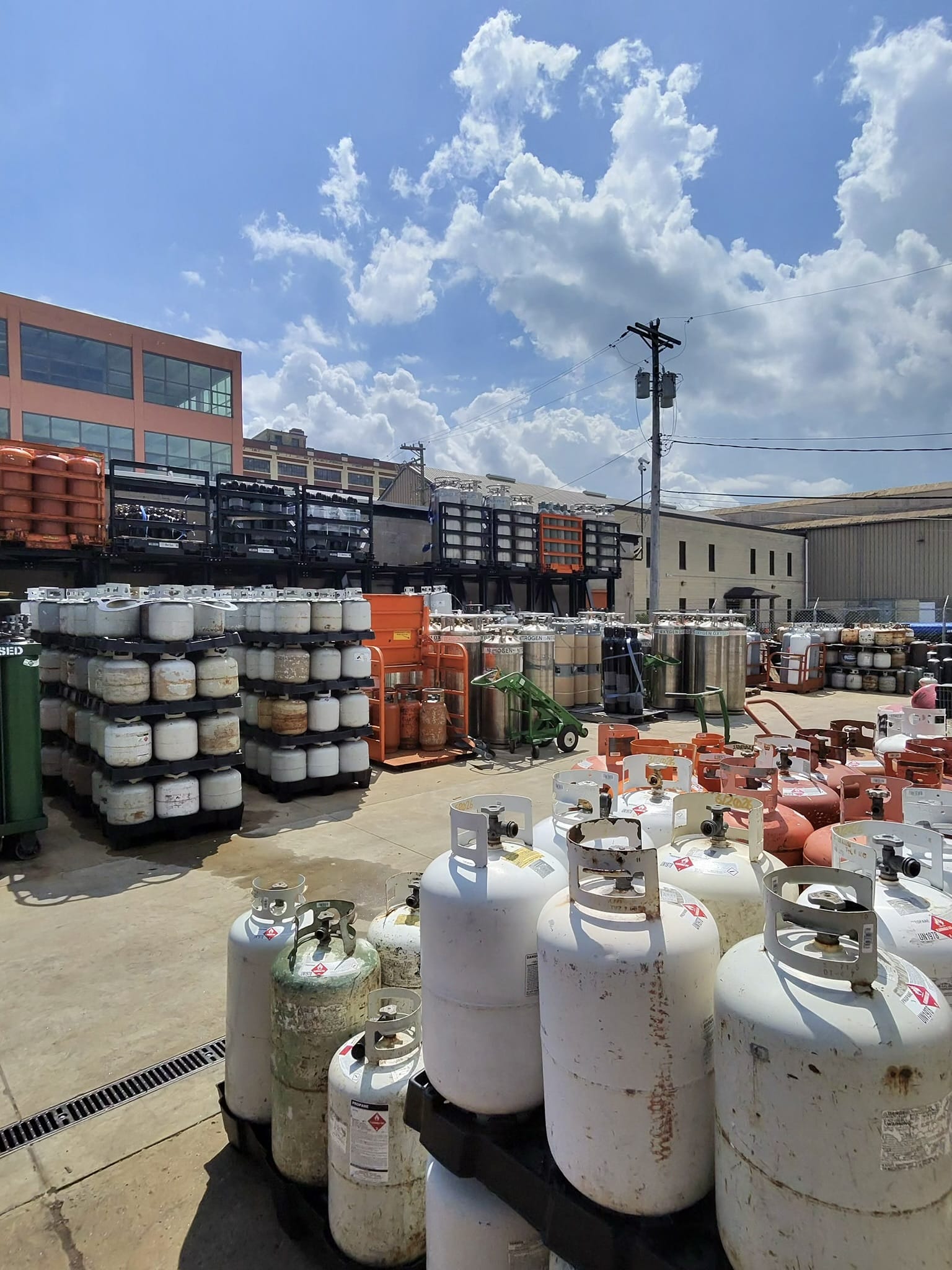 Outdoor industrial yard with stacked gas cylinders and propane tanks on pallets, power lines overhead, and a pink-brick building in the background.