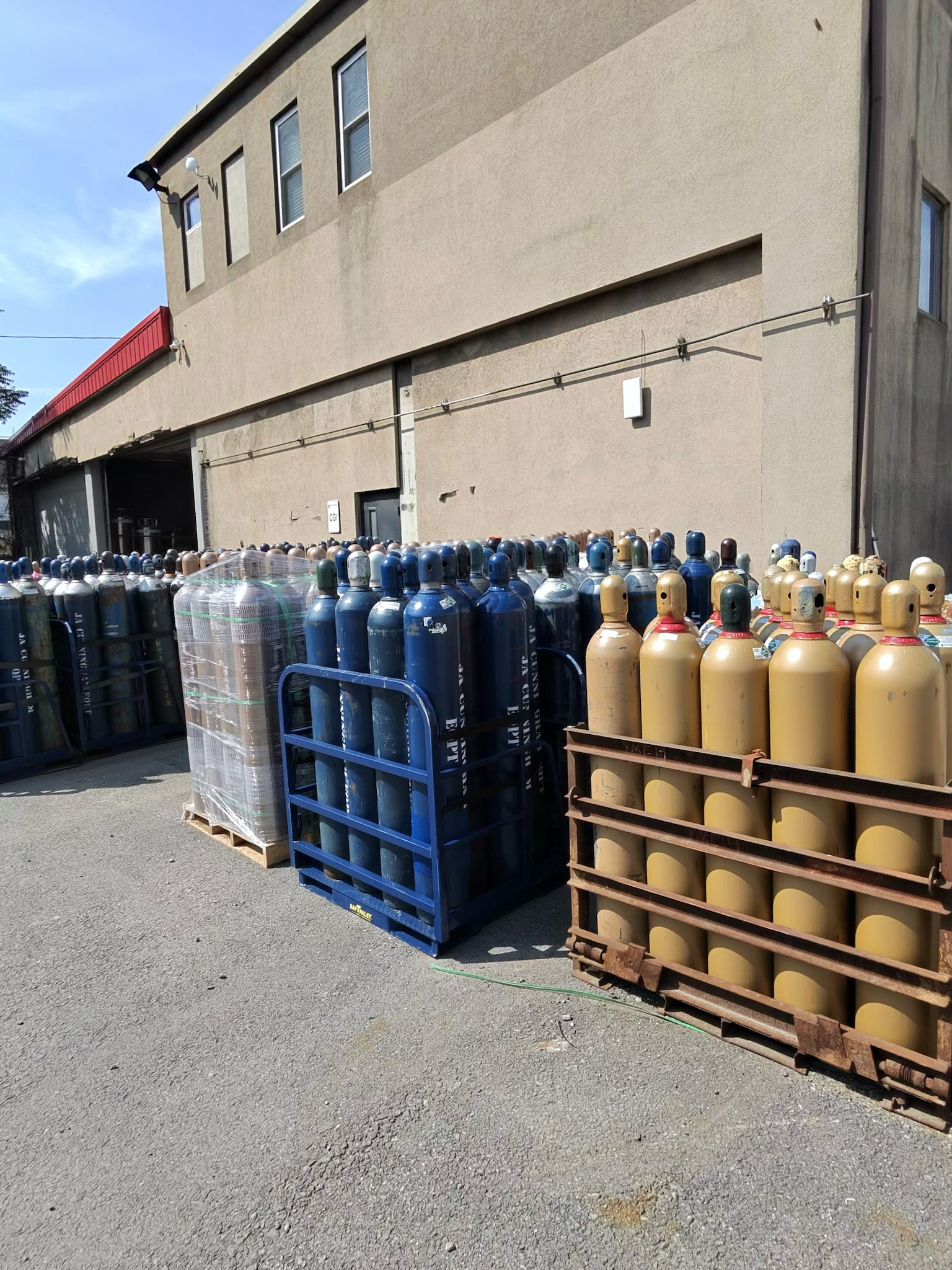 Stacks of blue and yellow compressed gas cylinders on pallets in an outdoor industrial yard beside a beige building with a loading bay.