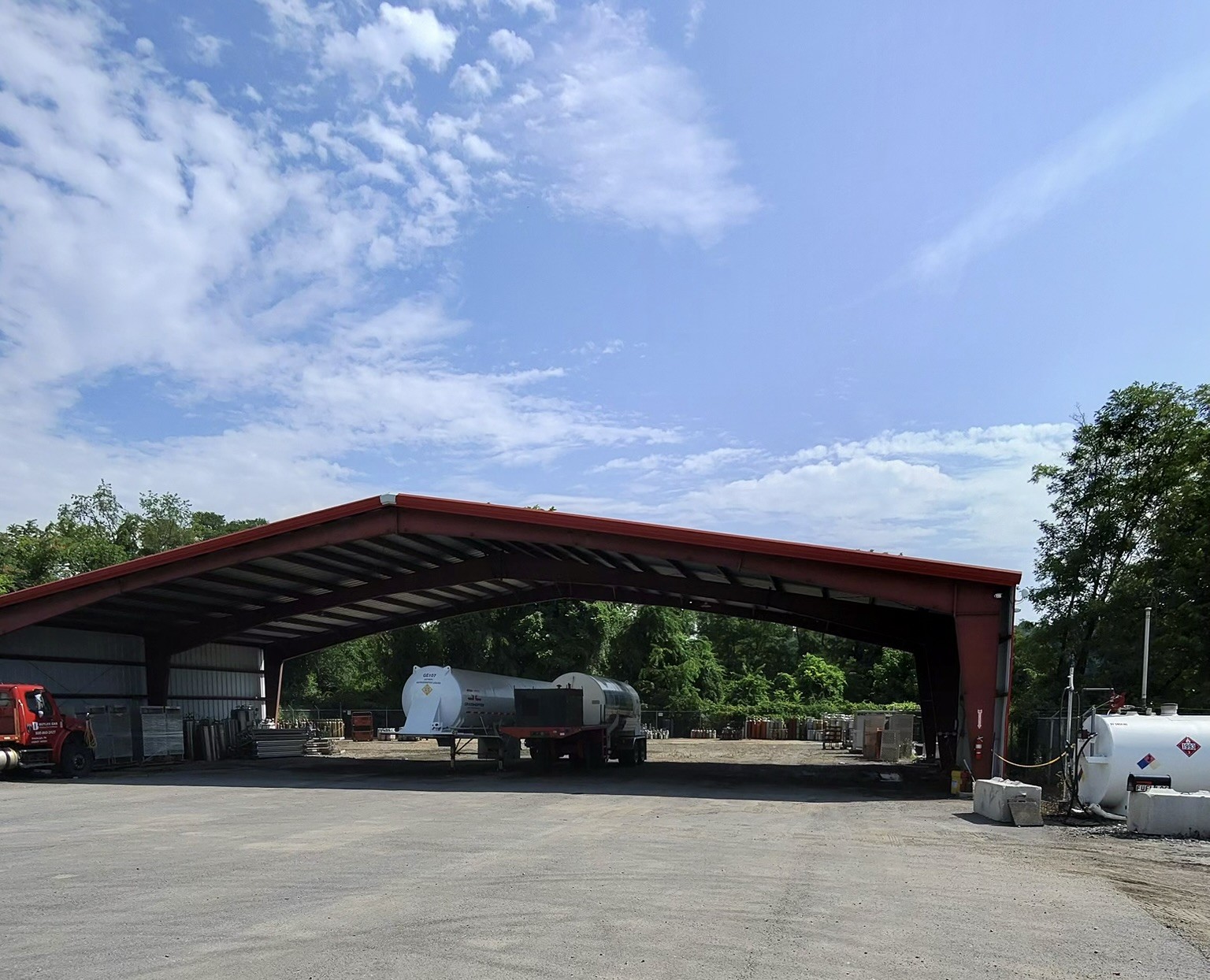 Open-sided red-roofed warehouse in an industrial yard, with tanks and equipment under the shelter and a truck nearby, under a blue sky.