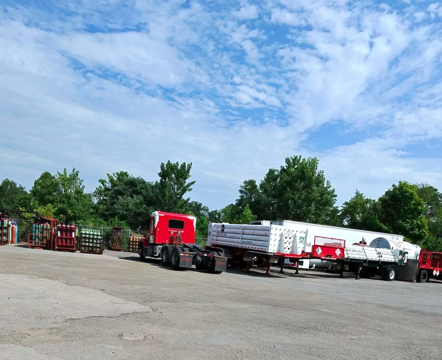 Red semi-truck with a white tanker trailered rig at a storage yard with gas cylinders and equipment on the left under a blue sky.