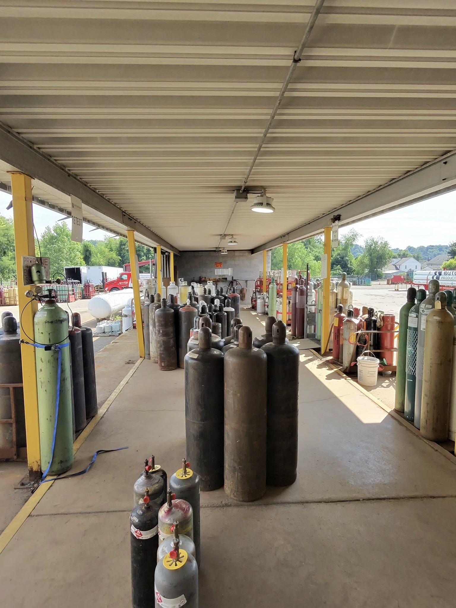 Open-air industrial storage area with rows of gas cylinders under a metal roof supported by yellow posts, outdoors near a warehouse complex.