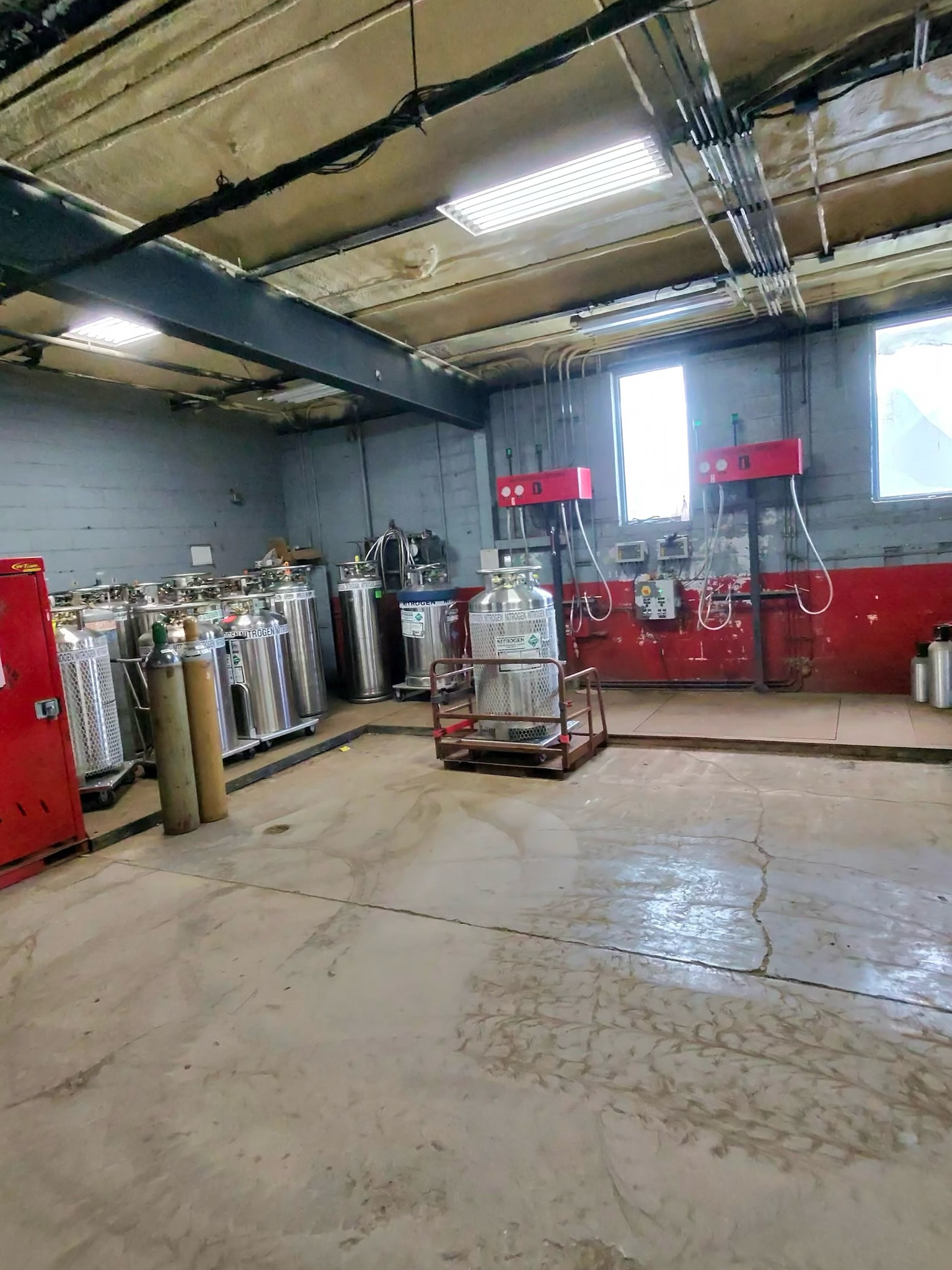 Industrial utility room with stainless gas cylinders, red control panels, and exposed pipes along a concrete floor.